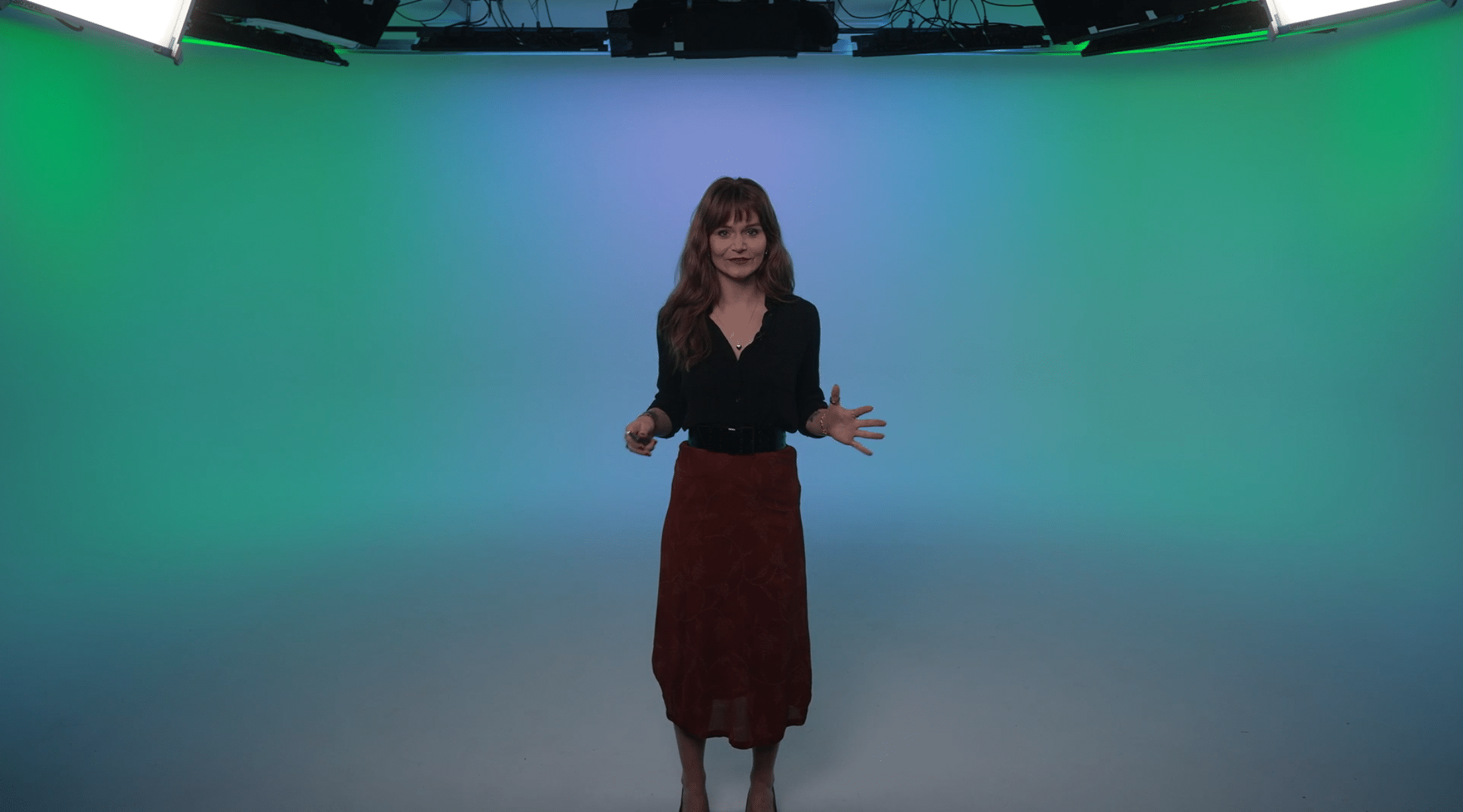 A woman stands in the center of a studio with a green and blue gradient background. She wears a black long-sleeve blouse and a red skirt, gesturing with her hands while smiling at the camera.
